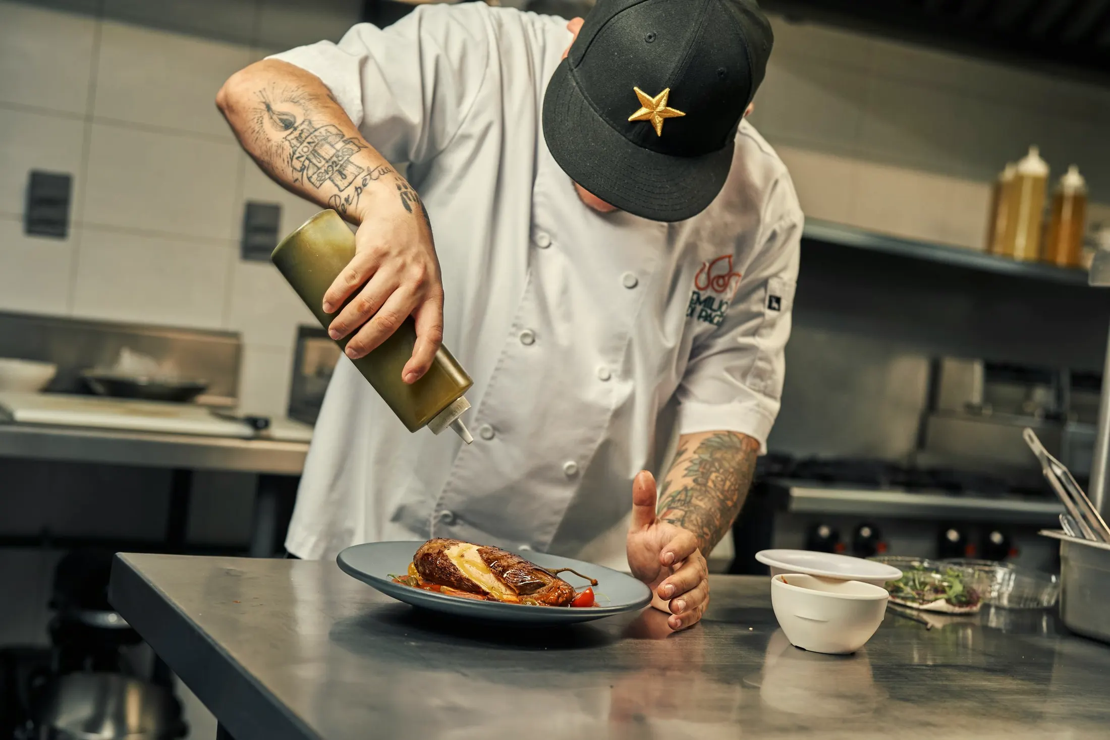 A chef in whites finishing a plated dish at a steel counter