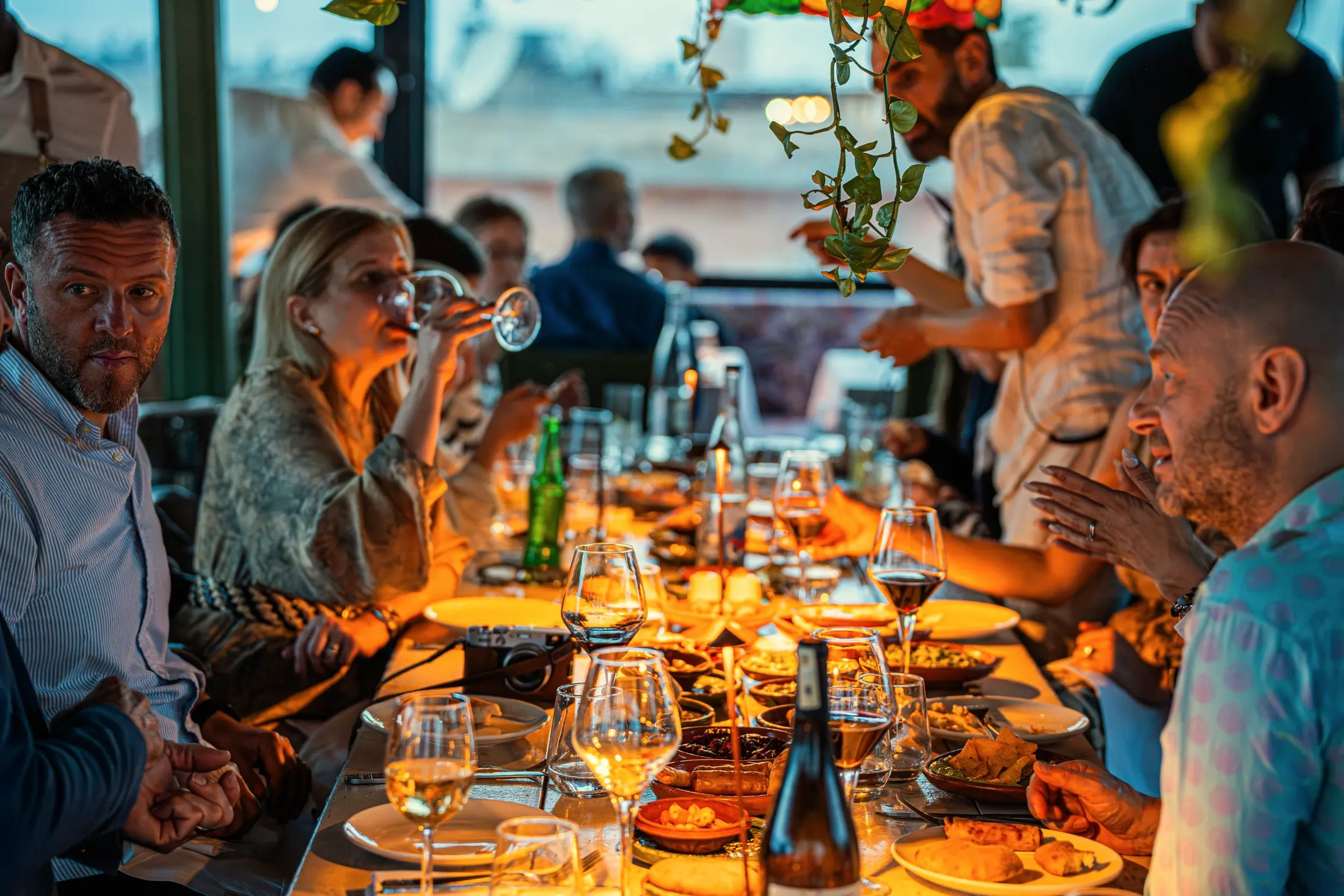 Friends around a long candlelit table at dinner, with wine glasses and shared plates
