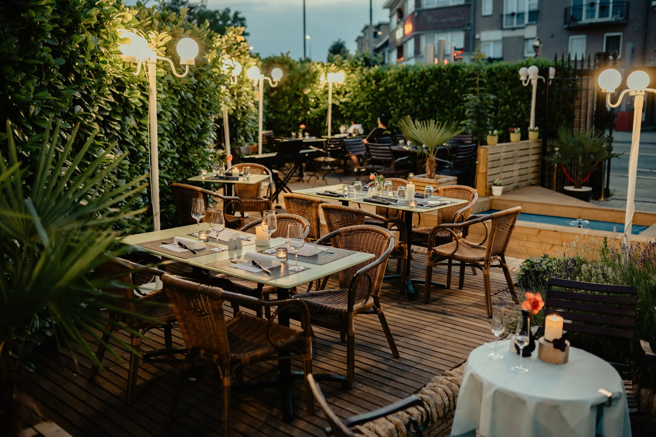 A restaurant terrace at dusk with wicker chairs, set tables and glowing lamps